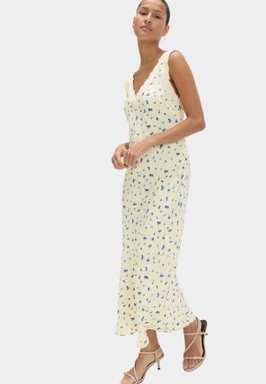 Kvinde iført en ærmeløs cremefarvet maxikjole med blå blomsterprint og blonder, kombineret med beige sandaler med hæl og stropper.