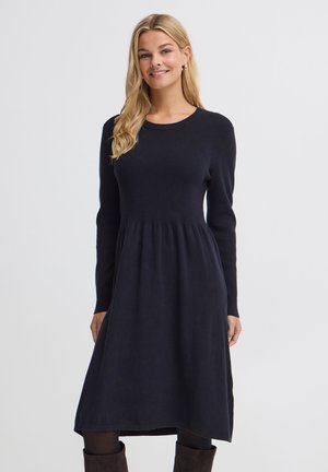 Femme aux longs cheveux blonds portant une robe noire à manches longues et des collants noirs, souriante sur un fond clair uni.