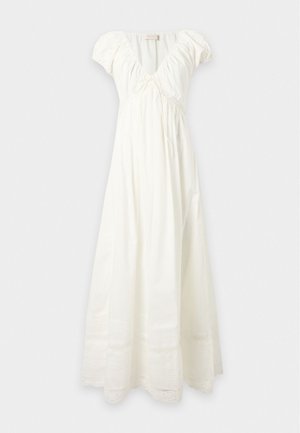 Femme portant une robe blanche à manches courtes et longueur cheville avec des détails en dentelle, debout, une main sur la hanche, chaussée de ballerines beige pointues.