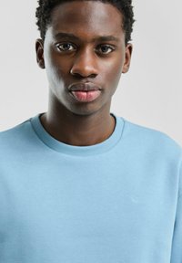 Jasnoniebieska bluza z okrągłym dekoltem, ściągaczami u mankietów i subtelnym logo QS na piersi. Tkanina wydaje się miękka i gładka.