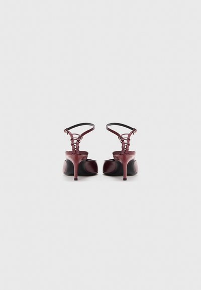 Par visokih tamnocrvenih potpetica s remenčićima za gležanj, snimljenih s leđa na jednostavnoj bijeloj pozadini.