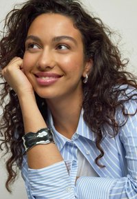 Rannekoru kiiltävässä hopeassa aaltoilevalla muotoilulla; mallilla on päällään vaaleansininen ja valkoinen raidallinen paita. Ihon sävy näyttää lämpimältä, ja hiukset ovat teksturoidut kiharat.