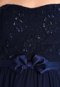 Mörkblå klänning med spetsok och paljetter, samt en satinmidja med detalj av en rosett. Flödande kjoldesign fulländar klänningen.