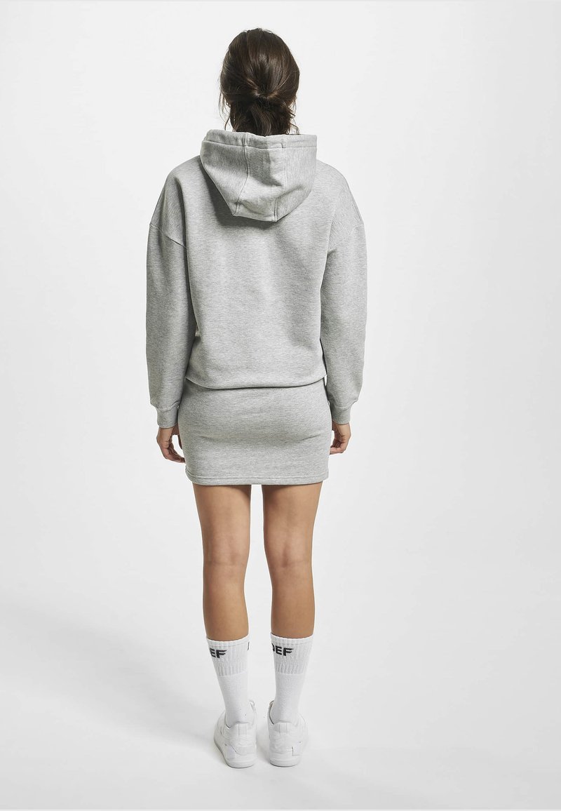 Femme aux cheveux foncés attachés, portant une robe sweat à capuche grise, des chaussettes blanches avec un texte noir et des baskets blanches, debout devant un fond blanc uni.