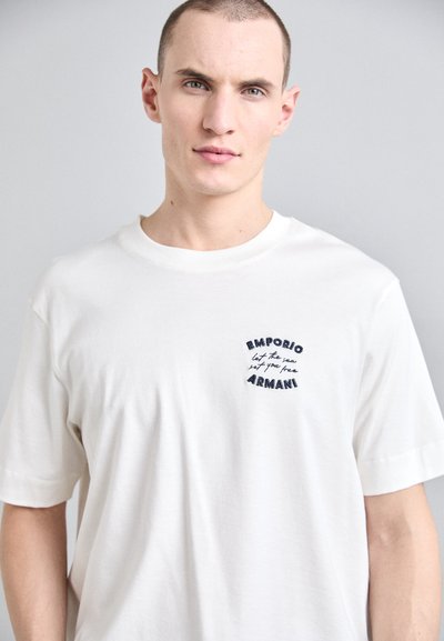 Valkoinen puuvillaiset t-paita lyhyillä hihoilla. Siinä on tummansininen brodeerattu logo: "Emporio Armani" ja lause "anna meren vapauttaa sinut."