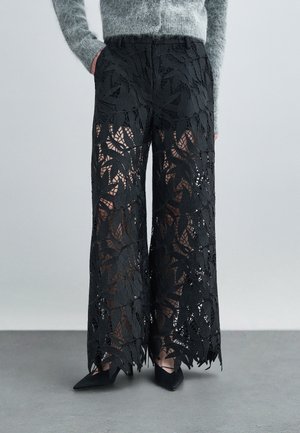 Pantaloni largi din dantelă neagră cu modele de frunze, parțial transparenți, având o talie elastică și margini detaliate. Asortați cu pantofi negri.