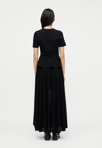 Γυναίκα με μακριά σκούρα μαλλιά που φοράει μαύρο κοντομάνικο μπλουζάκι και μαύρη μακριά φούστα που ρέει, στέκεται γυρισμένη προς τα πίσω σε απλό λευκό φόντο.