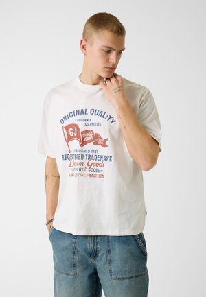 Młody mężczyzna w białej koszulce z grafiką w czerwonych i niebieskich napisach oraz niebieskich dżinsach, patrzący w dół z ręką przy brodzie.