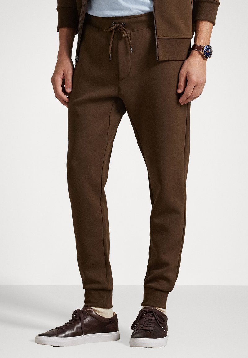 Pantalones de chándal marrones con cintura elástica, cordón ajustable, bolsillos laterales y puños acanalados. Combinados con zapatillas casuales oscuras.