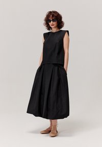 Czarny top bez rękawów w połączeniu z długą czarną spódnicą z plisami i kieszeniami. Zestawiony z płaskimi plecionymi butami i ciemnymi okularami przeciwsłonecznymi.