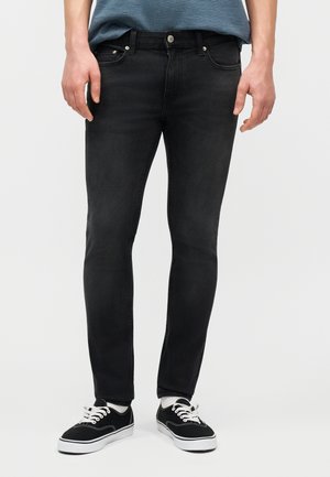 Mann iført slanke, svarte jeans, svart-hvite joggesko og en blåstripet t-skjorte, stående mot en ensfarget bakgrunn.