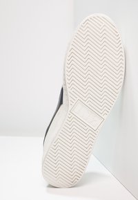 Svart sneaker med en vit gummisål, som har ett fiskbensmönster och texturerade detaljer. Logotypen syns på sidan av sålen.