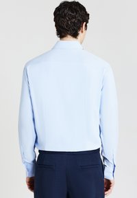 Mann mit dunklem, lockigem Haar, der ein hellblaues Langarmhemd in eine dunkelblauen Hose gesteckt trägt und mit dem Rücken zu einer neutralen Hintergrund steht.