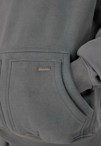Grauer Sweatshirt-Taschen mit einem kleinen Metalletikett "Dropsie" und einer Hand, die teilweise in die rechte Seite der Tasche eingeführt ist.