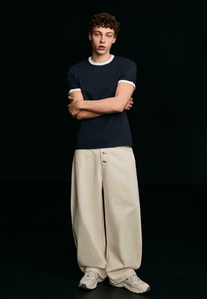 Mladý muž s kudrnatými vlasy stojí se založenýma rukama, má na sobě tmavě modré tričko s bílým lemováním, široké béžové kalhoty a bílé tenisky na černém pozadí.