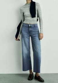 Top gris de manga larga, jeans de tiro alto en azul claro con pierna recta, terminado con mocasines marrones oscuros y un suéter azul marino colgado sobre el hombro.