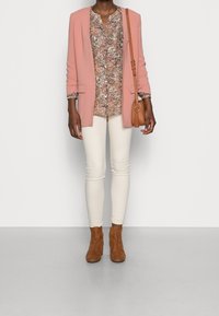 Blazer rose, blouse à fleurs dans les tons de rose et marron, skinny jeans crème, et bottines à talons marron. Sac en cuir à bandoulière couleur tan.