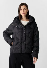 Kobieta w czarnej pikowanej kurtce puchowej z kapturem, stojąca z jedną ręką w kieszeni na jednolitym jasnym tle.