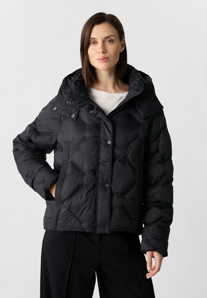 Kobieta w czarnej pikowanej kurtce puchowej z kapturem, stojąca z jedną ręką w kieszeni na jednolitym jasnym tle.