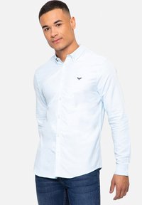 Camisa de manga larga azul claro con cuello, botonadura frontal y logo bordado en el pecho; combinada con jeans azul oscuro.