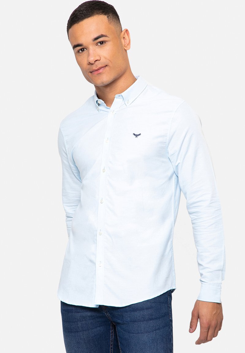 Camisa de manga larga azul claro con cuello, botonadura frontal y logo bordado en el pecho; combinada con jeans azul oscuro.