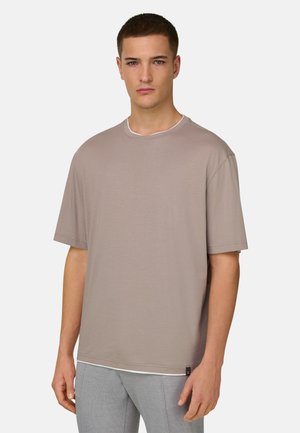 Ung mand med kort hår iført en løs beige t-shirt og lysegrå bukser, stående mod en ensfarvet lys baggrund.