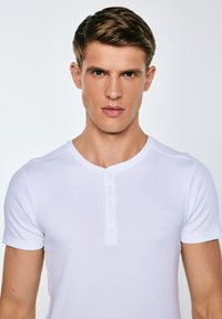 Fiatal férfi rövid barna hajjal, fehér rövid ujjú henley-ingben, szemben egy egyszerű háttér előtt.