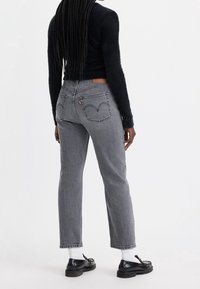Jeans de mezclilla gris con corte recto, cuentan con bolsillos traseros, parche con el logo de Levi's y una textura ligeramente desgastada. Llevados con una blusa negra y zapatos.