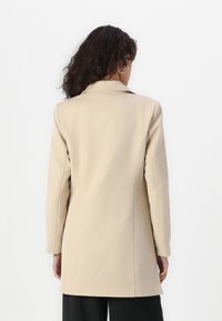 Manteau beige avec un col structuré, des manches longues et une coupe ajustée. Tissu lisse avec une texture subtile, atteignant une longueur mi-cuisse.