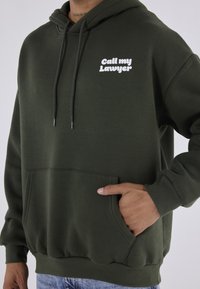 Pehmeästä kankaasta valmistettu oliivinvihreä huppari, jossa on kengurutasku, kiristysnauhallinen huppu ja valkoinen tekstitys "Soita asianajajalleni" rinnan kohdalla.