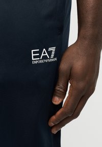 Σκούρο μπλε αθλητικό σορτς με το λογότυπο EA7 Emporio Armani τυπωμένο σε λευκό χρώμα. Κοντινό πλάνο που δείχνει ένα χέρι και την υφή του υφάσματος.