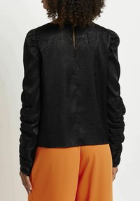 Blusa nera testurizzata con maniche a sbuffo e dettaglio a goccia sul retro, abbinata a pantaloni arancioni, che evidenziano una finitura liscia.