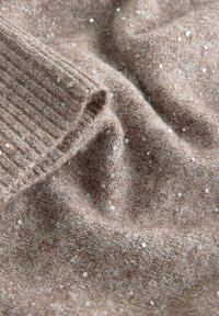 Soft beige knitted fabric with small shiny sequins scattered evenly across its textured surface and ribbed edge detail.