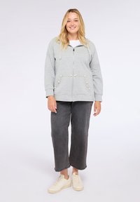Felpa grigia con zip e dettagli decorativi di perline nelle tasche, vestibilità comoda, abbinata a pantaloni grigi larghi e sneakers beige chiaro.