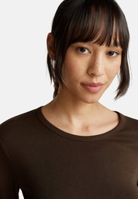 Jonge vrouw met steil donker haar en een pony, gekleed in een bruine ronde hals shirt, die naar voren kijkt met een neutrale uitdrukking.