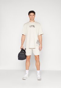Beige bomullströja med grafisk tryck, kombinerad med beige shorts. Håller en svart duffelväska. Vita strumpor och lätta sneakers fulländar outfiten.