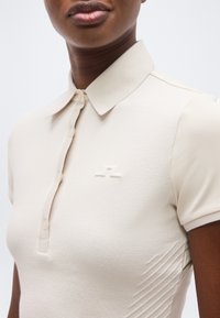 Κοντινό πλάνο ενός ατόμου που φοράει μπεζ πόλο μπλουζάκι με κοντό μανίκι, με ένα μικρό λογότυπο σε τονοποιημένα χρώματα στο στήθος και μια κουμπωτή λαιμόκοψη.