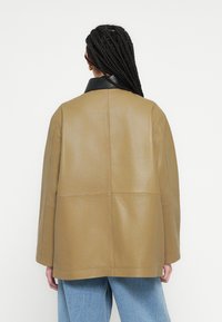 Tādas krāsas ādas jaka ar melnu apkakli, ar vaļīgu piegriezumu un minimālām šuvju detaļām, redzama no aizmugures.