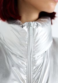 Silberne, metallische Puffjacke mit hohem Kragen, strukturierten Falten und einem zentralen Reißverschluss mit strukturiertem Finish.