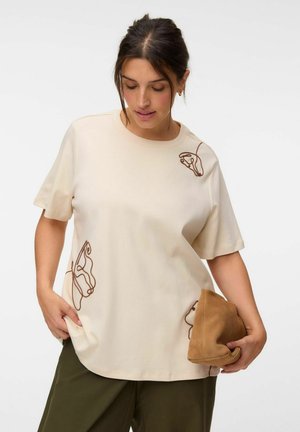Femme aux cheveux foncés porte un t-shirt crème avec des motifs abstraits de lignes marron, un pantalon olive, et tient un sac en suede marron plié.