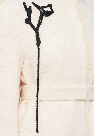 Šviesiai rožinė vilnonė striukė su tekstūruota taškų raštu, turinti juodą pynimo užsegimą ir paprastą audinio diržą.