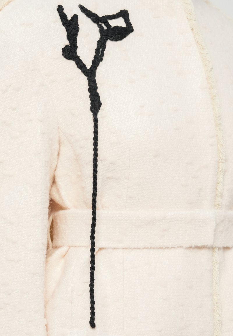 Giacca di lana rosa chiaro con motivo a pois testurizzati, dotata di una chiusura con laccio intrecciato nero e una cintura in tessuto semplice.