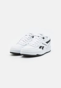 Tenisi din piele albă cu accente negre, având un vârf perforat, șireturi plate și o talpă din cauciuc texturată. Logo-ul Reebok pe lateral.