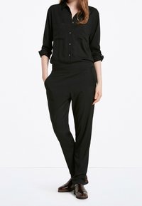 Zwarte long sleeve shirt met knopen aan de voorkant en twee borstzakken, gecombineerd met zwarte tapse broek. Beide gemaakt van soepele stof. Bruine schoenen.