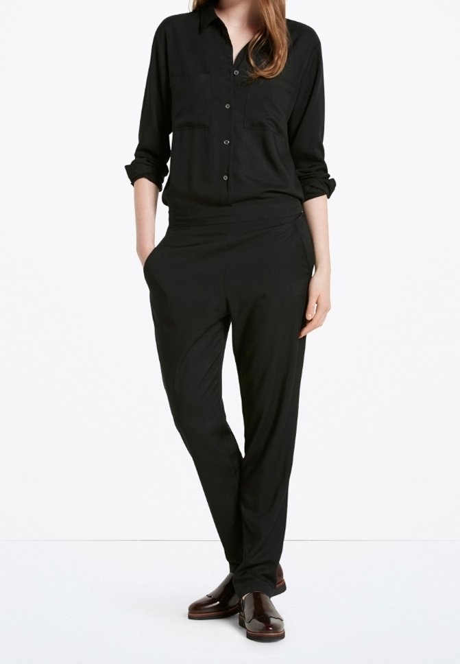 Zwarte long sleeve shirt met knopen aan de voorkant en twee borstzakken, gecombineerd met zwarte tapse broek. Beide gemaakt van soepele stof. Bruine schoenen.