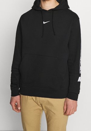 Homme portant un sweat à capuche Nike noir avec logo blanc sur la poitrine et la manche, associé à un pantalon beige, debout devant un fond uni.