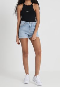 Bodysuit negro con cuello halter y texto "OBEY", combinado con pantalones cortos de denim azul claro y zapatillas blancas. El fondo es blanco plano.