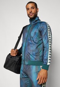 Chaqueta de chándal con cremallera en degradado verde y azul, con franjas laterales que presentan detalles de logotipo. Acompañada de un bolso de viaje negro con detalles de malla.