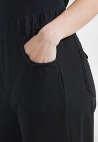 Pantaloni de trening negri din material moale, cu talie elastică și buzunar lateral cu margini rotunjite. Textură netedă și croială casual.