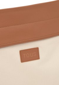 Beżowa tkanina pleciona w woreczku z brązowym skórzanym akcentem, wyposażona w zamknięcie na suwak i tłoczoną naszywkę z logo. Gładka faktura z czystymi liniami.